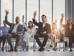 Group of businessmen and women sitting on chairs and all raising their hands in an office setting.