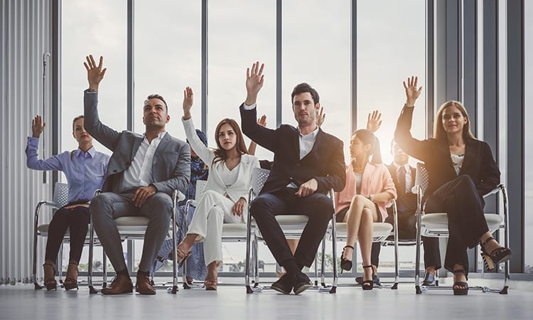 Group of businessmen and women sitting on chairs and all raising their hands in an office setting.