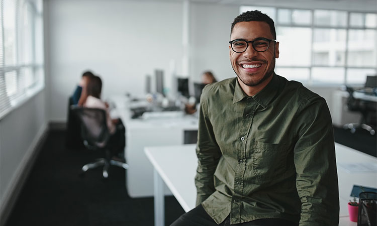 Man with glasses smiling while sitting on a desk in the office